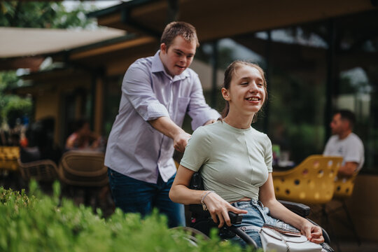 A boy with Down syndrome joyfully pushes a smiling girl in a wheelchair outside a cafe, showcasing friendship and support in a sunny, inviting environment.
