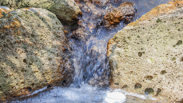 A natural scene of a crystal-clear stream flowing between weathered, porous rocks. The water cascades gently, creating foam, and the textured rocks display earth-toned colors