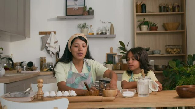 Medium shot of positive mother talking to little kid, showing all ingredients they need for making homemade pastry, girl learning and smiling at home kitchen