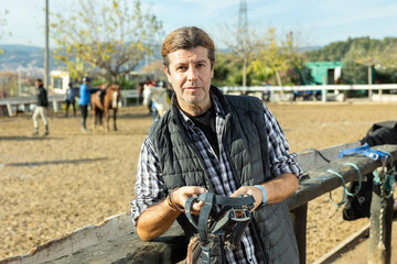 Middle-aged man instructor holding horse halter or reins during working day at horse club on sunny...