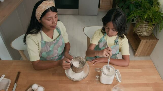 Top view shot of Latin aunt in hairband and apron sitting at table with small niece and showing her how to sift flour with sifting cup for making batter in modern home kitchen