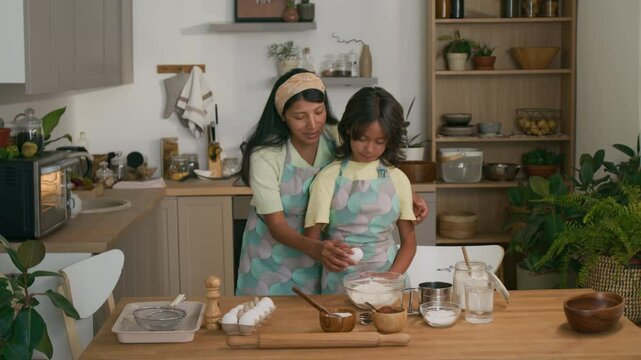 Full medium shot of calm Hispanic mother in apron teaching her little daughter to make batter, she cracking egg over flour in glass bowl while standing by table in modern kitchen