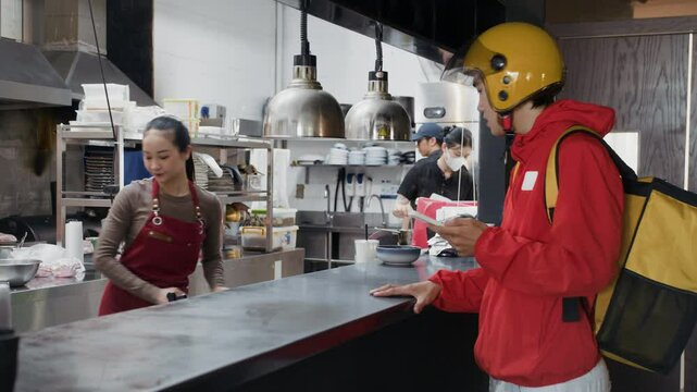 Medium long shot of deliveryman wearing yellow helmet and red jacket receiving takeout bag with food from female restaurant worker inside kitchen