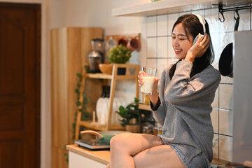 Happy young woman sitting in a kitchen with a glass of milk and enjoying music on headphones