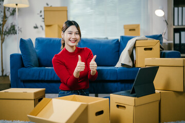 Young woman is smiling and giving thumbs up while unpacking boxes in her new apartment. She is...