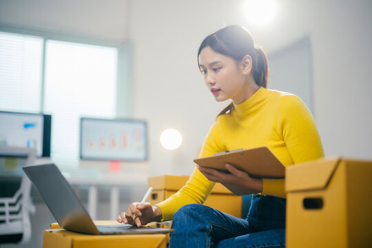 Manager is working on a laptop and checking orders on a clipboard while sitting surrounded by cardboard boxes in a warehouse