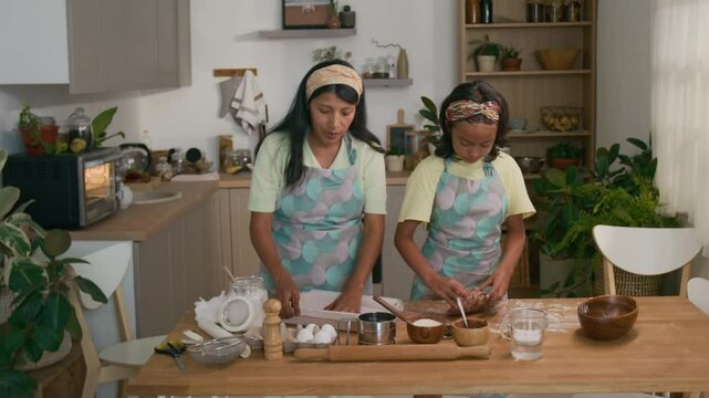 Full medium shot of little Hispanic girl kneading dough and helping mom who preparing baking tray for cookies, both wearing hairbands and aprons and cooking together in modern kitchen