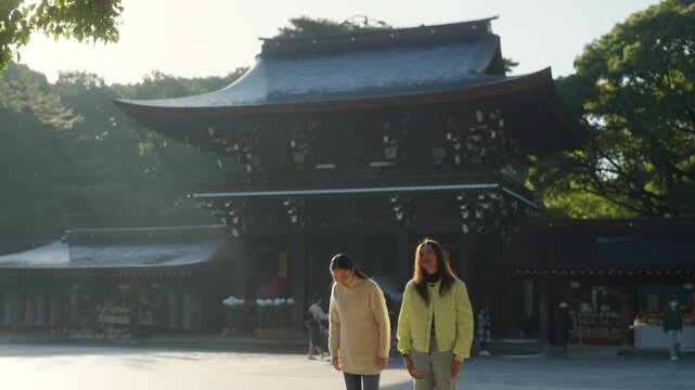 Two Asian woman friends praying in traditional Meiji Jingu Shrine in Tokyo city, Japan in autumn. People tourist travel Japan landmark famous place and learning Japanese culture on holiday vacation.