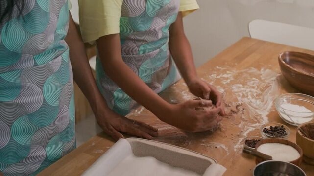 Side midsection shot of anonymous child in apron learning how to knead dough, adding chocolate drops into it, making homemade cookies together with mother in kitchen