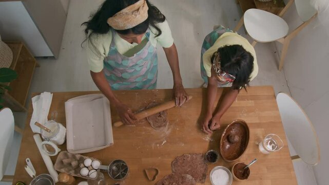 Top view of woman showing how to roll out dough and passing rolling pin to daughter, teaching her to make homemade pastry in home kitchen