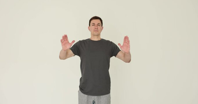 Concentrated man in t-shirt raises hands to show namaste gesture on white background. Middle aged male practices yoga exercises and mindfulness