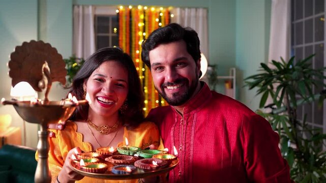 A beautiful Indian couple celebrates Diwali night by lighting Samai and oil diyas,holding laddoos in hand,sharing love,joy and happiness under the glow of festive lights