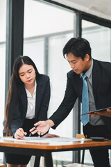 Two business professionals in formal attire discussing project details in a modern office with large windows and natural light.