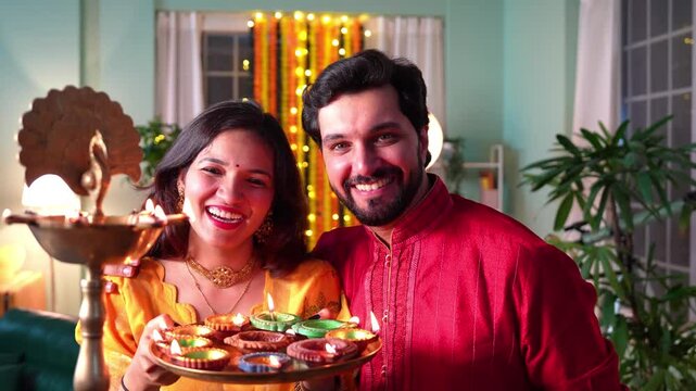 A beautiful happy Indian couple lights Samai and adorns home with colourful painted diyas,enjoying delicious laddoos in a joyous Diwali celebration.