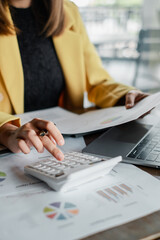 Professional woman in yellow blazer using a calculator and reviewing financial documents at a desk with a laptop.
