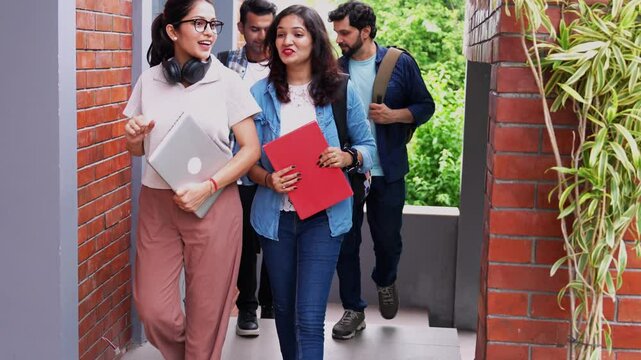 Indian Asian college friends happily walking together down a busy corridor, carrying bags, books, and a laptop, excitedly chatting about their classes and plans for the day ahead