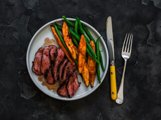 Medium-rare beef steak, sweet potato, string beans - a delicious lunch on a dark background, top view