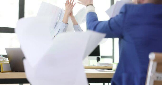 Businesspeople joyfully throw papers in air clapping hands in office. Colleagues express happiness over finishing difficult commercial project
