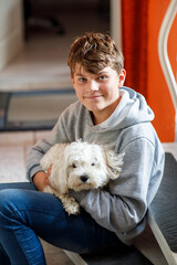 Happy teenager boy and cute maltese puppy together. Handsome child holding his dog at home, hugging. Pet love, family dog.