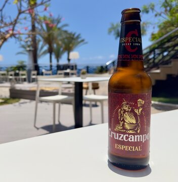 Tenerife, Spain - May 29, 2024: A closeup of a bottle of Cruzcampo Especial beer on a table against a defocused outdoor seating area in the background. 