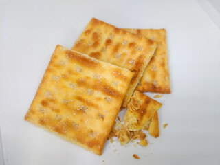 A few crispy crackers, one partially eaten, lie on a white surface with visible crumbs. The image showcases the flaky texture, golden color, and sugar crystals, emphasizing a snack in mid-bite.