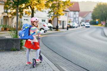 Little happy girl on way to school by pushing scooter. Elementary school child riding in the city, with big satchel. Kid with pink helmet. Safe route to school and movement for children concept.