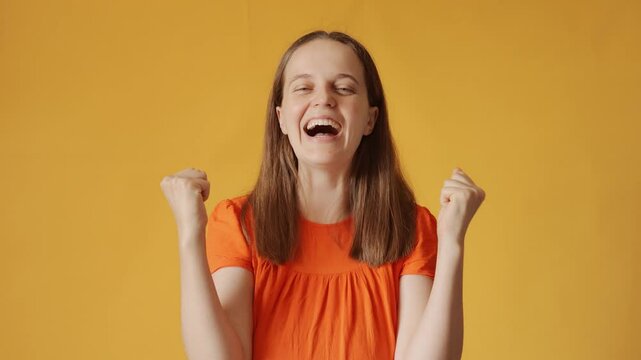 Excited woman with brown hair wearing orange shirt celebrating her success with raised hands and joyful shout expressing happiness and triumph celebrating victory isolated over yellow background