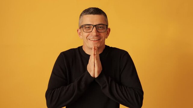 Joyful man with glasses wearing black raglan celebrating victory and success with hands together in excitement expressing happiness and satisfaction isolated over yellow background