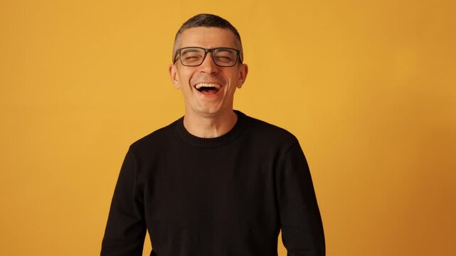 Joyful man wearing black raglan and glasses laughing out loud with toothy smile enjoying funny moment and expressing happiness isolated over yellow background