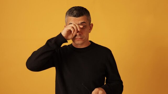 Unwell man wearing glasses and black raglan showing fatigue and pain in eyes while taking off spectacles and rubbing eye grimacing isolated over yellow background