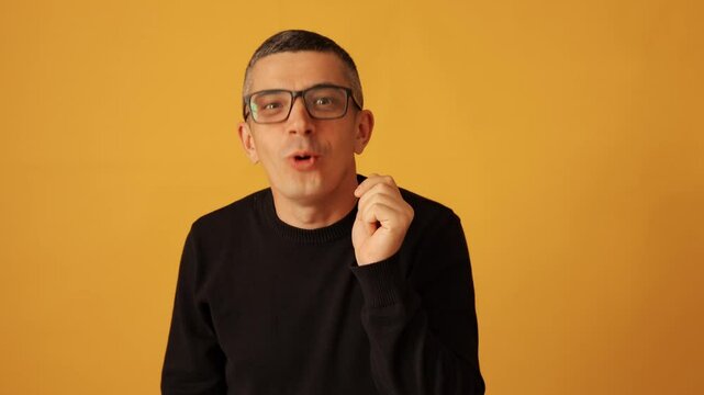 Curious man wearing black raglan and glasses listening intently with hand raised to ear focused on conversation and expressing interest isolated over yellow background
