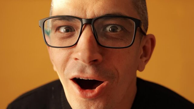 Closeup of amazed man in glasses wearing black raglan jumper expressing shock with open mouth and big eyes reacting to unexpected news isolated over yellow background
