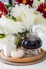 Still-life. White and red dahlia flowers in a vase, a teapot, a pumpkin and a house on a white table in the interior of a home kitchen. A cozy autumn concept.