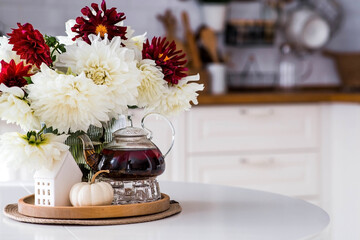 Still-life. White and red dahlia flowers in a vase, a teapot, a pumpkin and a house on a white table in the interior of a home kitchen. A cozy autumn concept.