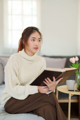 Young asian women in sweater reading a book to thinking and learning about literature story while sitting on couch to relaxation and doing activity to spending time with slow life lifestyle at home