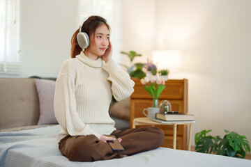 Young asian women in sweater wear headphone and listen music with enjoying on smartphone while sitting on the couch to relaxation and doing activity for spending time with slow life lifestyle at home