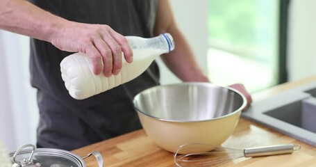 Chef takes bottle with milk pouring ingredient into bowl at wooden countertop. Man adds dairy ingredient following traditional recipe slow motion