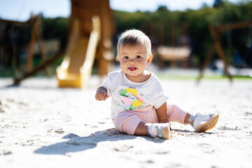 Adorable baby sitting on sandy playground in sunlight