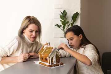 mother and daughter decorating gingerbread house. Beautiful living room with lights. Happy family celebrating holiday together.