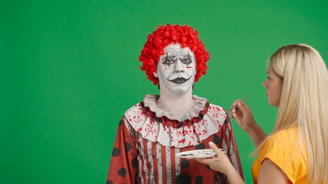 Scary halloween man dressed as clown standing in studio, woman stylist finishing spooky makeup, painting male face, isolated on chroma key green screen background.