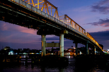 The Kapuas River Bridge in Pontianak glows beautifully at sunset, reflecting warm lights on the water. The scene captures the serene transition from day to night, showcasing urban charm.