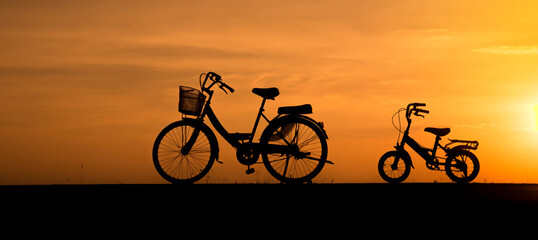 Silhouette of adult and child bicycles parked at sunset. Outdoor leisure and cycling of happy family activity moments concept with vibrant sky background-banner image.