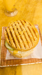 Grilled chicken cheeseburger on a yellow table with juice beside it, all set on a wooden surface. A vibrant, appetizing food scene.