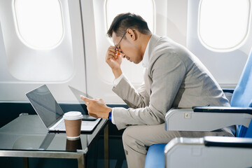 frustrated man sitting on an airplane with her head in her hands. Asian Businessman sitting in airplane working laptop and tablet