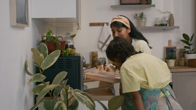 Medium shot of smiling Hispanic aunt taking tray of freshly baked chocolate cookies out of mini-oven and enjoying smell together with little niece, going towards table in modern kitchen