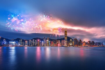A cityscape with brightly lit skyscrapers reflecting in the calm water below. Multicolored fireworks explode in the evening sky above. © YiuCheung