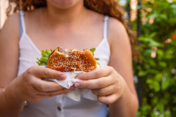 Closeup of young womans hands holding bitten juicy double cheeseburger with sesame seed bun, fresh lettuce and beef. Teenager girl is eating a burger wrapped in paper in fast food restaurant on summer