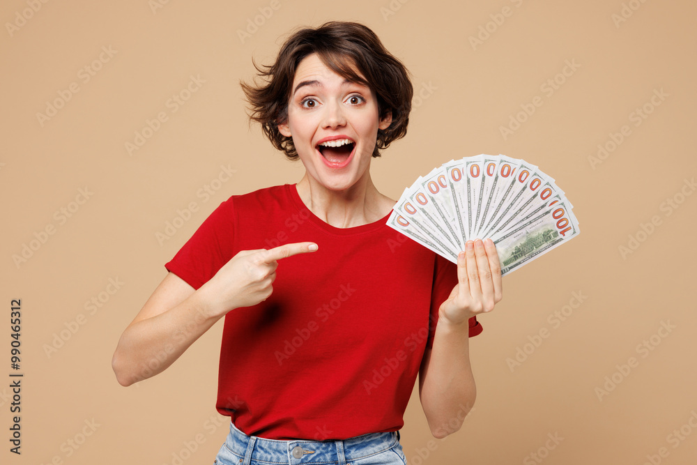 Wall mural Young smiling happy woman she wearing red t-shirt casual clothes hold in hand fan of cash money in dollar banknotes isolated on plain pastel light beige background studio portrait. Lifestyle concept.