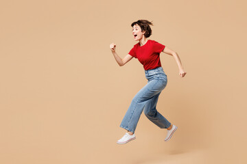 Full body side view excited young smiling happy woman she wear red t-shirt casual clothes jump high run fast hurry up isolated on plain pastel light beige background studio portrait Lifestyle concept