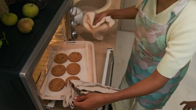 Top view of anonymous woman in apron taking hot tray with freshly baked chocolate chip cookies out of oven in modern kitchen, little child looking happy